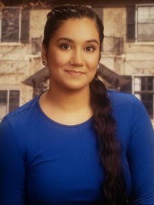 Headshot of Eshta Bhardwaj. Estha is seated in front of an older beige house. She has her hair over one shoulder and is wearing a blue long-sleeve shirt.