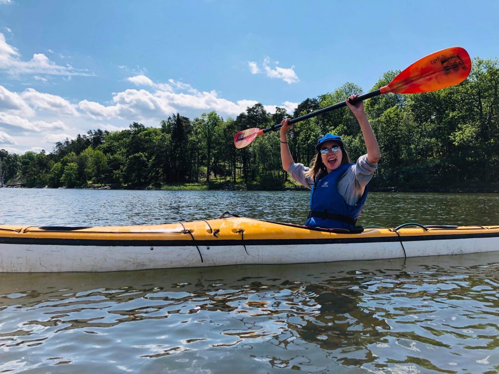 Ruby Lindsay in a yellow and white kayak holding her red and yellow paddle above her head. Ruby is on a body of water in a wooded area with trees in the background and a blue sky with some clouds. Ruby is wearing a blue baseball hat and sunglasses.