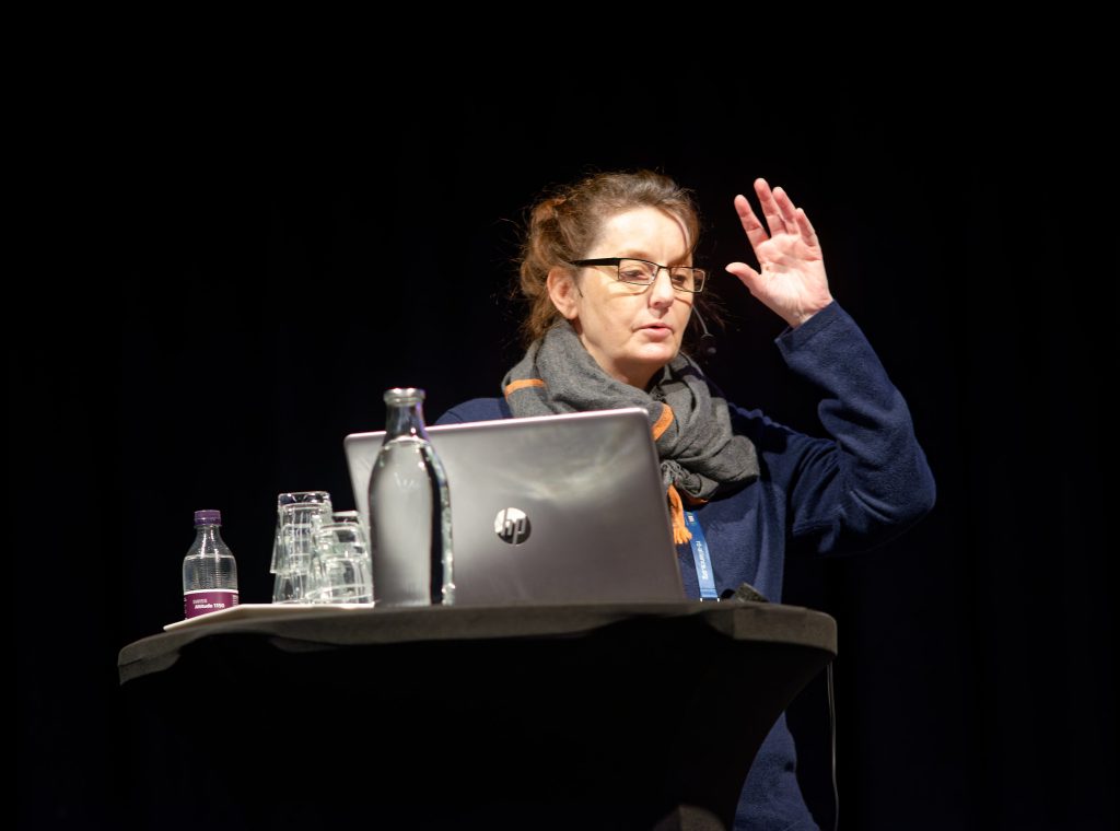 Karen Payne is standing at a podium and speaking, with her left hand raised to demonstrate a point.