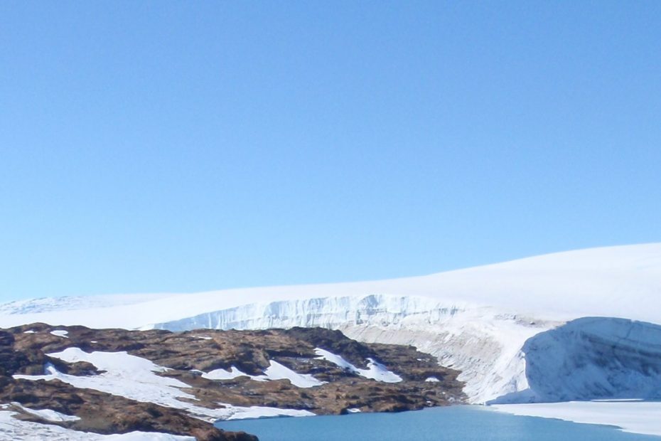 A frozen landscape with dry tundra on the left meeting a body of water that appears to be frozen towards to the right. A ice sheet is visible in the background, and sky is clear and blue.